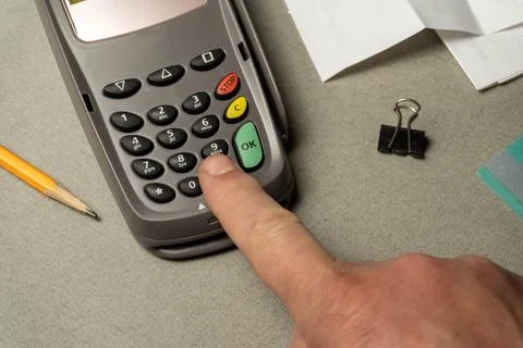 A finger presses the button on a payment device while office supplies like .. Stock Photos