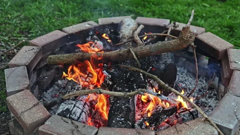 Fire burns in a circular stone pit as someone adds sticks in a backyard during Stock Footage 334159347