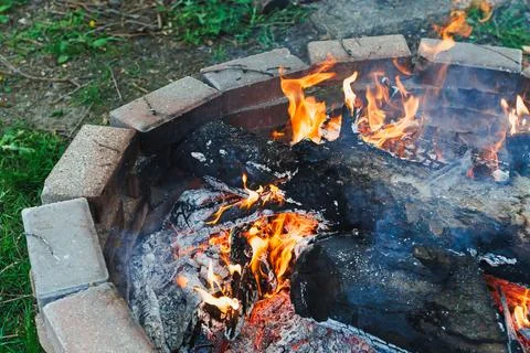 Fire burns in a circular stone pit in a backyard during evening time with l.. 스톡 사진