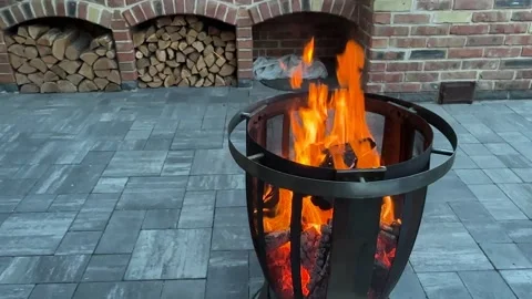 Fire burns in a metal pit on a stone patio in the evening with stacked wood in Stock Footage 339574877