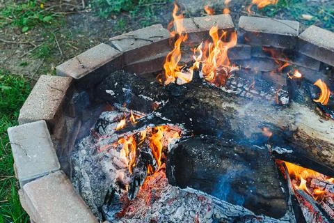 Fire burns in a stone fire pit surrounded by grass and wood during the even.. Photos