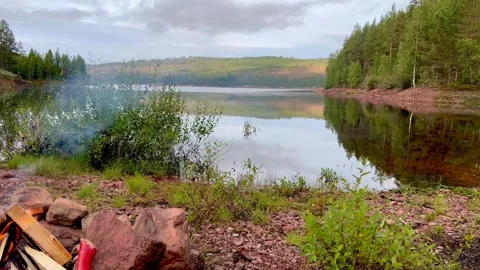 A fire lit on the beach overlooking a still lake and mountains Stock Footage 297666109