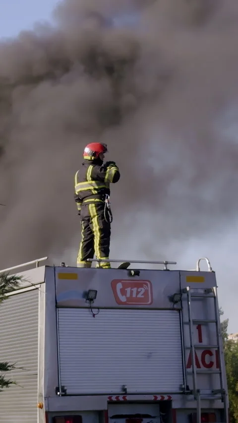 Firefighter in full gear is positioned on fire truck, combating intense flames Stock Footage 315445866