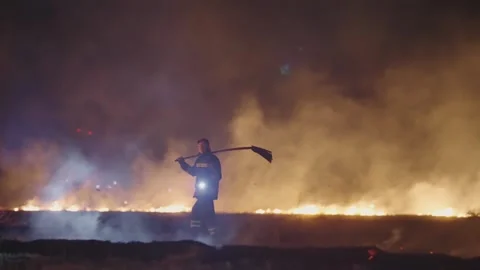 A firefighter walk in front of a large fire, holding a broom and light search Stock Footage 280012479