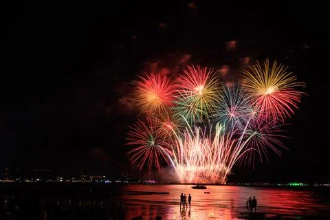 Fireworks Exploding And Illuminating Night Sky Over Ocean Beach With Crowd .. Stock Photos