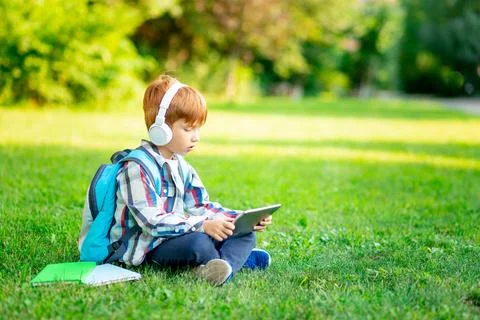 A first-grader boy with a backpack and a tablet with headphones on a green .. Photos