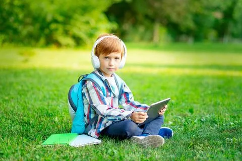 A first-grader boy with a backpack and a tablet with headphones on a green .. Photos