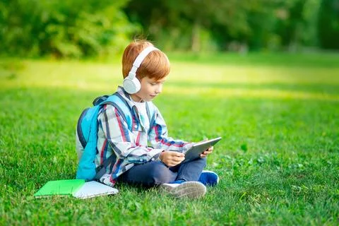 A first-grader boy with a backpack and a tablet with headphones on a green .. Stock Photos
