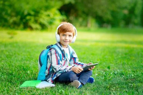 A first-grader boy with a backpack and a tablet with headphones on a green la Photos