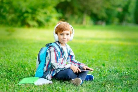 A first-grader boy with a backpack and a tablet with headphones on a green la 스톡 사진