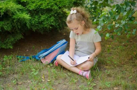 First grader sits having crossed legs under a tree and does homework. Stock Photos