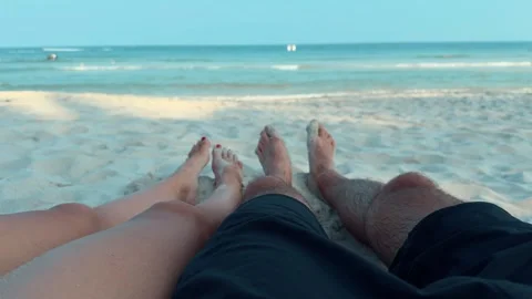 First-person view couple's legs rest on sandy beach looking at sea immersed in Stock Footage 266663570