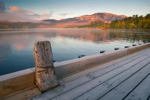 First sun rays hit mountain above the lake with wooden pier in foreground. Red Stock Photos