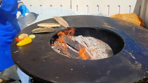 Fish and vegetables cooked on round cast-iron outdoor grill with fire in middle Stockbeeldmateriaal 197487326