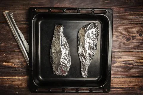 Fish wrapped in foil on a baking sheet and on a wooden table. Cooking fish in Stock Photos