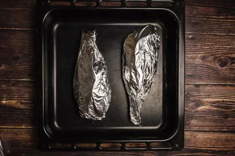 Fish wrapped in foil on a baking sheet and on a wooden table. Cooking fish in Stock Photos