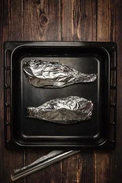Fish wrapped in foil on a baking sheet and on a wooden table. Cooking fish in Stock Photos