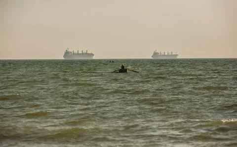 Fisherman in a boat between two cargo ships. Stock Photos