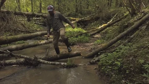 Fisherman in cap climbs over trees trunks in muggy stream Stock Footage 112538968