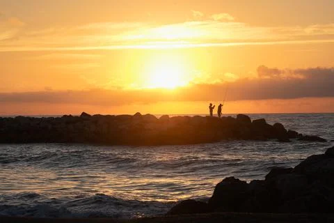 Fishermen on a pier in the light of the setting sun, surrounded by the sea .. Stock Photos