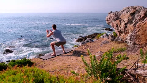 Fit barefoot man does jump squats on coastal cliff overlooking Atlantic ocean Stock Footage 323292175