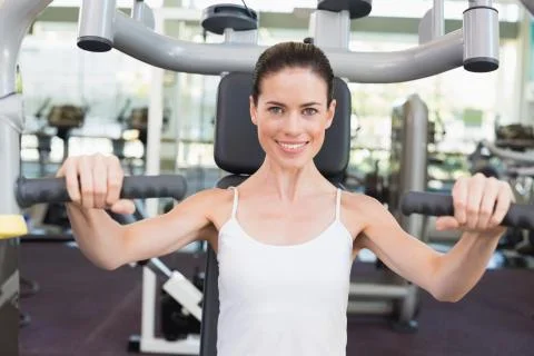 Fit brunette smiling at camera using weights machine for arms 스톡 사진