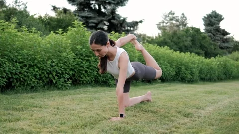 Fitness instructor practicing a balancing yoga pose on a grassy lawn in a public Stock Footage 295245251