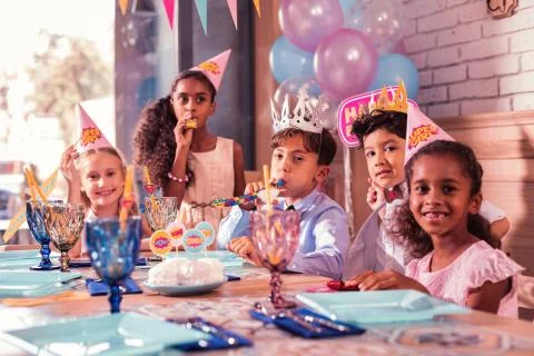 Five cute children sitting at the table and blowing party horns Foto stock