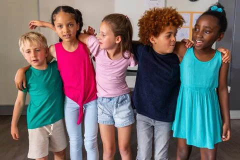 Five school age friends standing in line linking arms in classroom with artwork Stock Photos