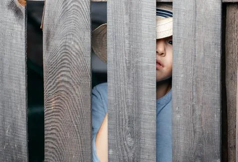 Five- to six-year-old kid in a straw hat peeps through a wooden village fence Stock Photos