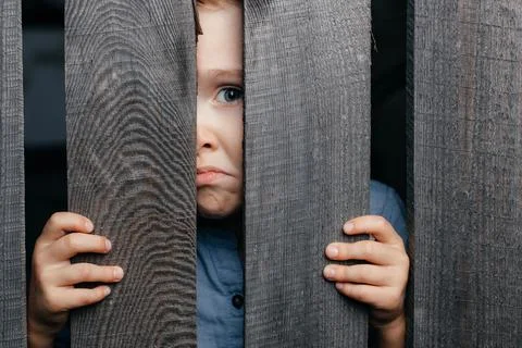 Five- to six-year-old kid in a straw hat peeps through a wooden village fence Stock Photos