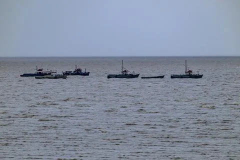 Five small boats float on choppy grey water. Cloudy sky looms over distant se Stock Photos
