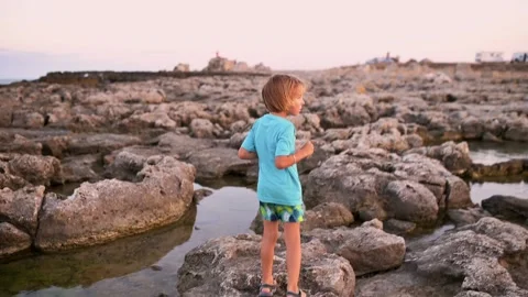 A five-year-old boy with long, light hair agilely leaps along the rocky seaside Video stock 250829117