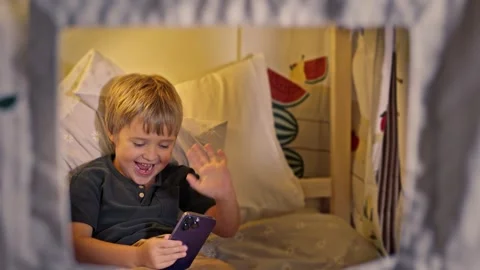 Five-Year-Old Boy Saying Hello with a Smartphone in Hand, Lying on Bed at Home Stock Footage 279531852