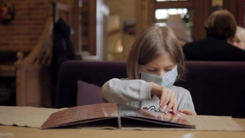 A five-year-old girl in a protective face mask studies the menu while sitting at Video stock 146096239