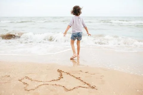 Five years cute curly caucasian girl painting on sand on the beach. Stock Photos