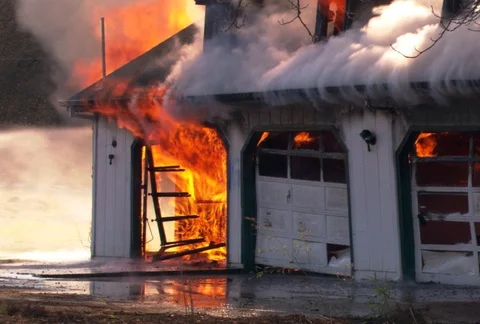 Flames and steam curl up from under the eaves of a blazing garage while an Vidéo 86774227