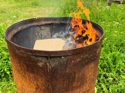 Flames dance vigorously within a rusty barrel, surrounded by lush green grass Stock Photos