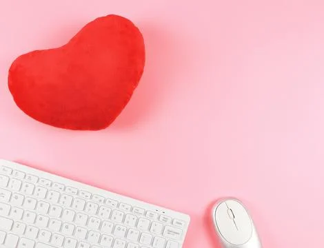 Flat lay of computer keyboard and mouse with red heart shape pillow on pink.. Stock Photos