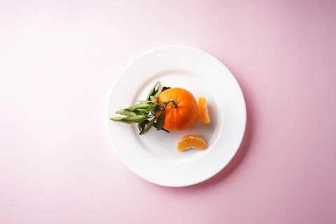 Flat lay mandarin with a green branch and slices in a white plate on a pink b Stock Photos