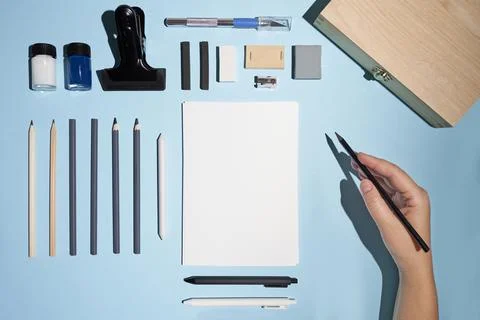 Flat lay notepad, pencils and other art staff with hand holding pencil on blue. Stock Photos