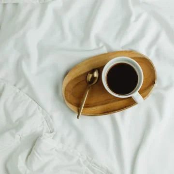Flatlay of cup of coffee and spoon on wooden tray in bed on white sheets, mor Foto stock