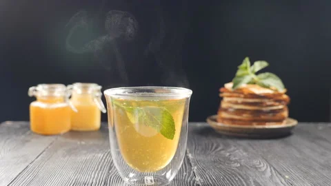 Flavored tea is steaming on black background with honey and stack of pancakes in Stock Footage 133135348
