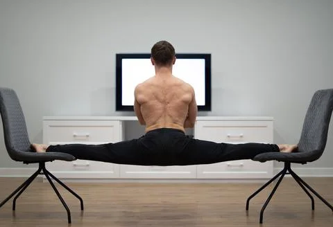 Flexible man keep balance in split between two chairs watching TV. Yoga and Stock Photos