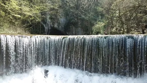 Flight above small waterfall trough suns flares toward the spring in side cav Stock Footage 106158467