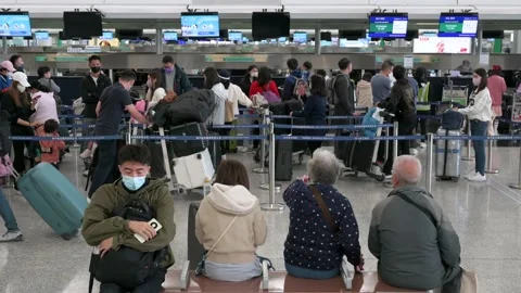 Flight passengers sit on a bench as other travelers check in at the Stock Footage 235652690