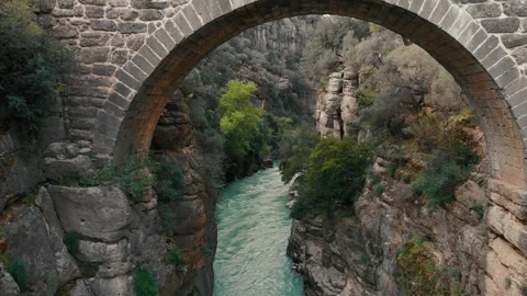 Flight under stone road bridge above water flowing in wild creek between cliffs Stock Footage 247508683