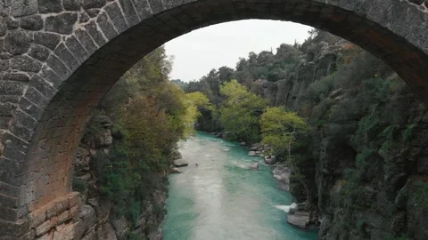 Flight under stone road bridge above water flowing in wild creek between cliffs Stock Footage 248461616