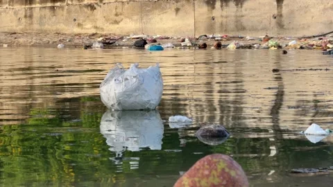 A floating plastic bag drifts through a polluted river, surrounded by scattered Vídeo Stock 325431917
