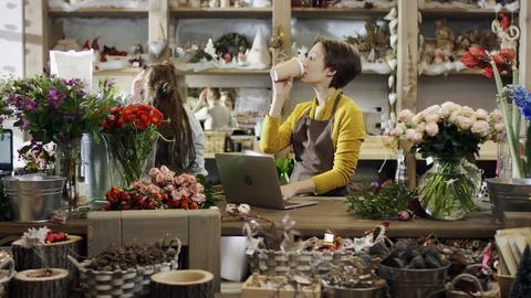 Florist in yellow blouse prints something on a laptop, holding the cup in her Stock Footage 101704689